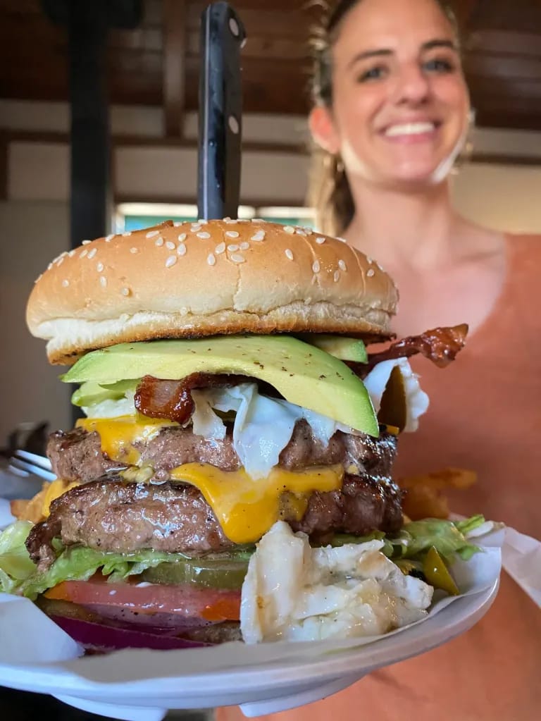 A double cheeseburger stacked with bacon, avocado and jalapeño, a knife driven through it, carried out to a table