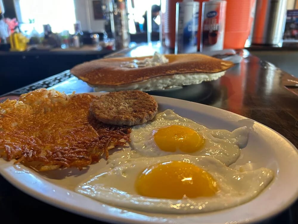 Two fried eggs, hash browns and a sausage patty on a plate, a pancake on the counter behind