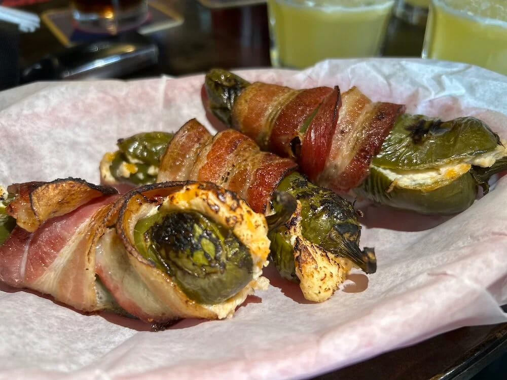 Bacon-wrapped jalapeños stuffed with cream cheese in a paper-lined basket, margaritas behind them