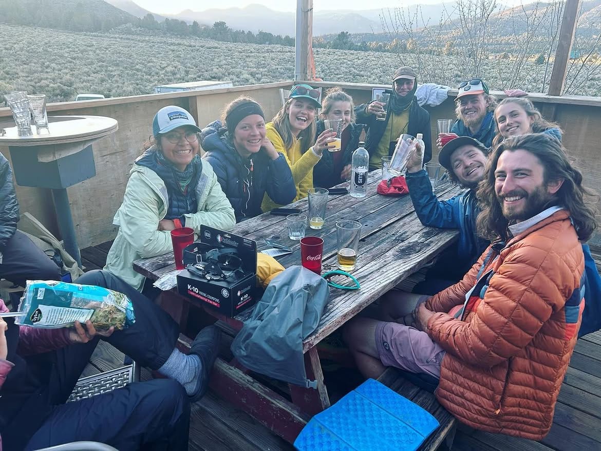 A long picnic table of hikers at dusk, packs and bottles across the boards