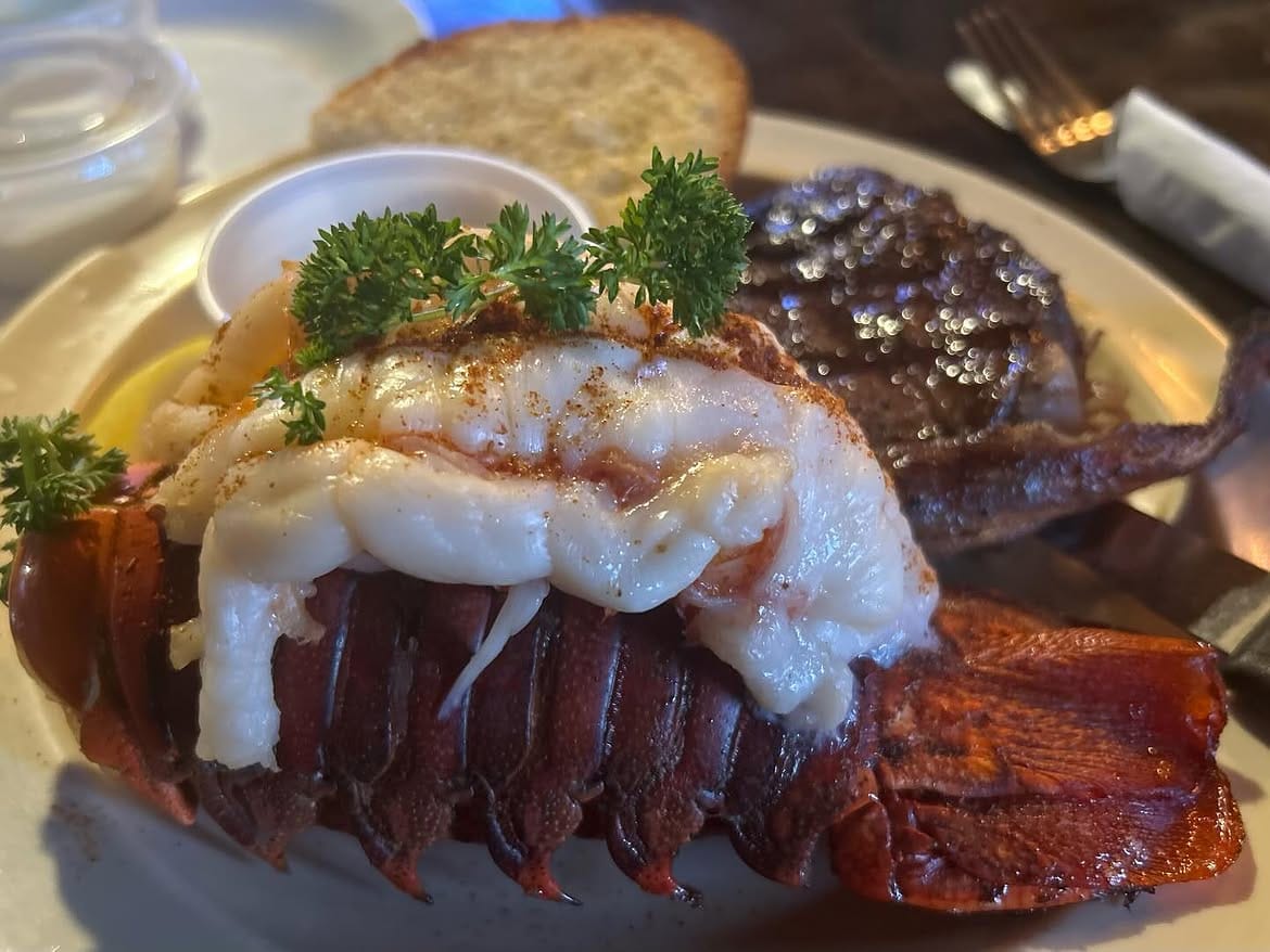 A whole lobster tail plated beside a seared steak, garnished with parsley and served with garlic bread