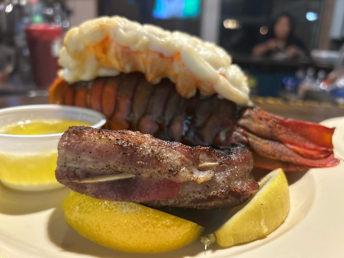 A close-up of lobster tail and peppered steak on a plate with lemon wedges and a cup of drawn butter