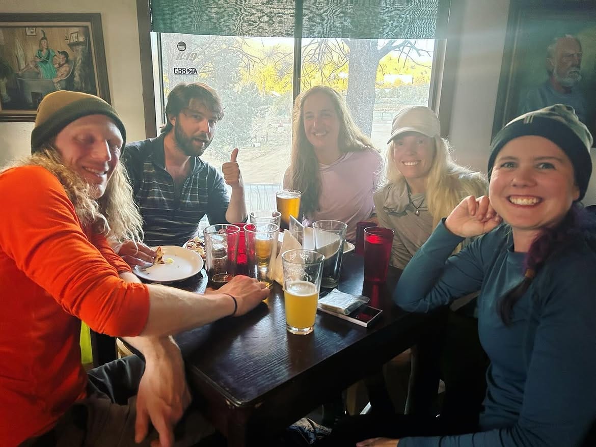 Hikers around a table by the window, plates and glasses between them